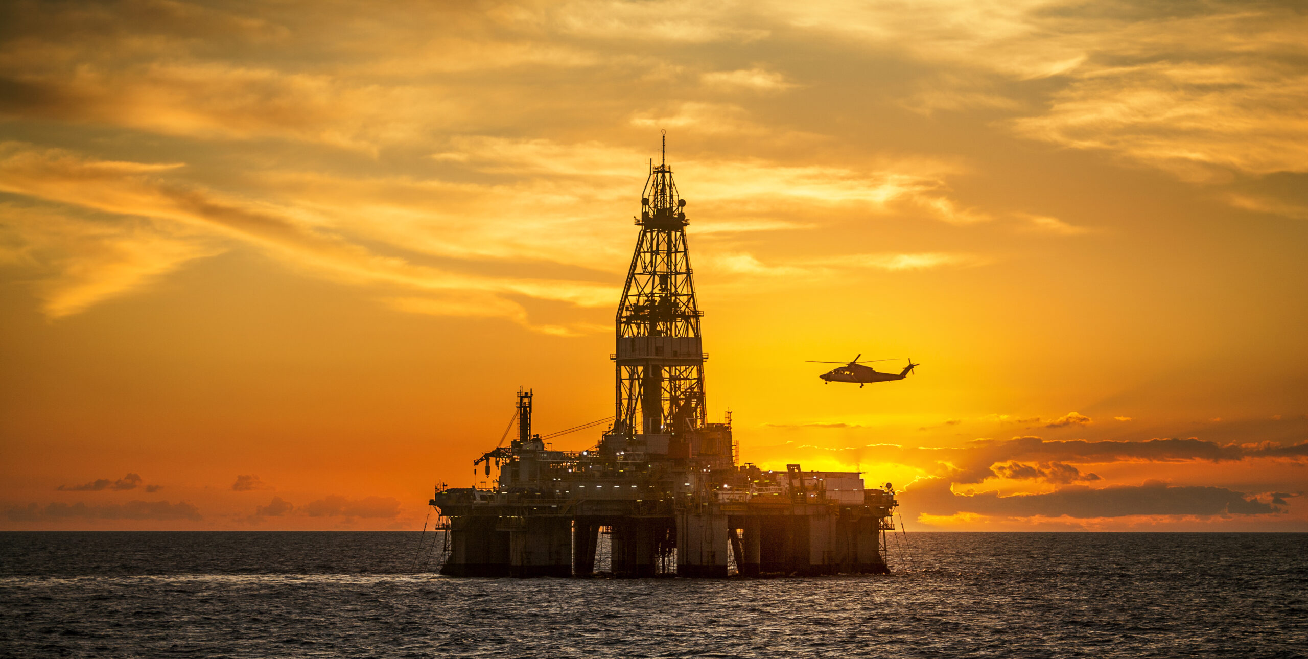 A helicopter flies over an offshore oil rig surrounded by water at sunset.