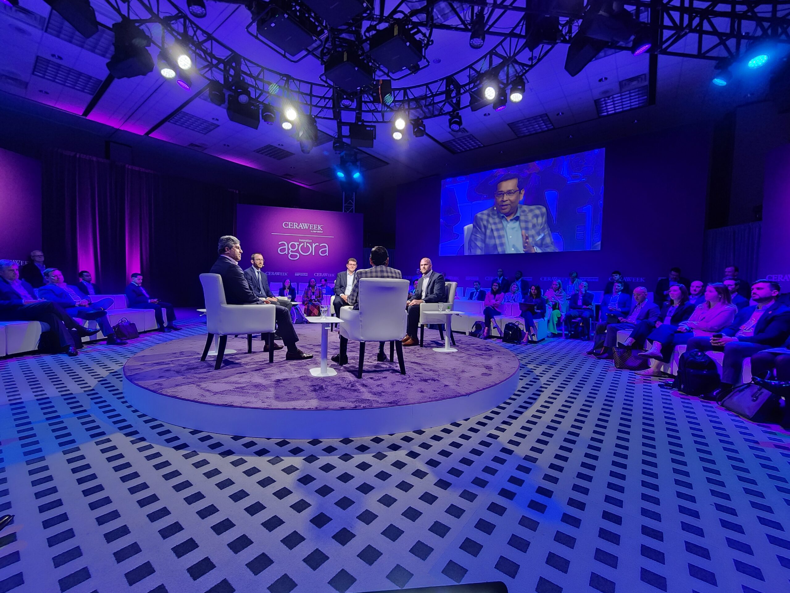 Four people sit in a circle in the middle of a conference hall as people look on. 