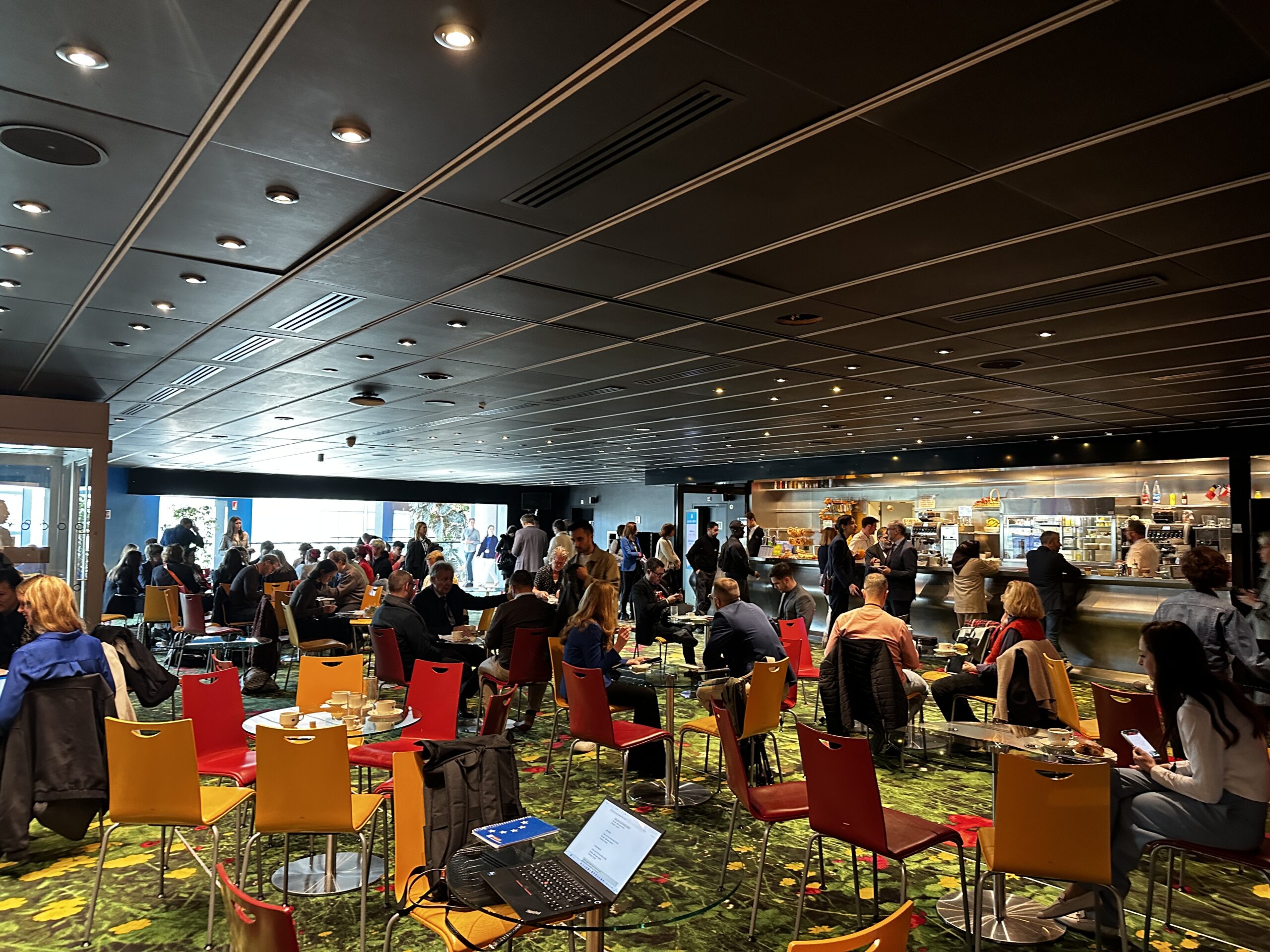 Inside a crowded bar in the EU plenary building. Round tables are surrounded by colorful chairs, many of them with people sitting in them. 