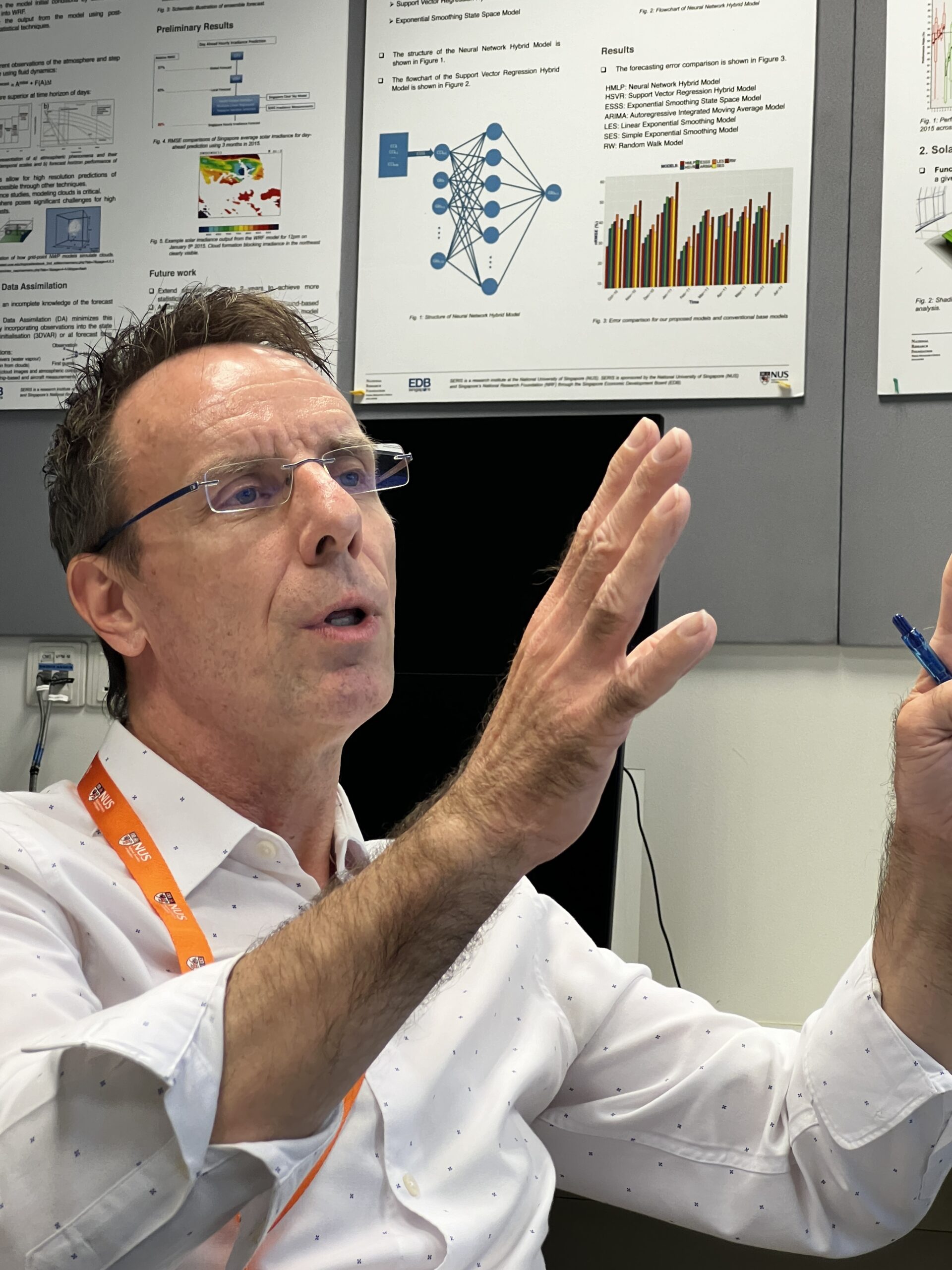 A man gestures while speaking, sitting in an office in front of some graphs.