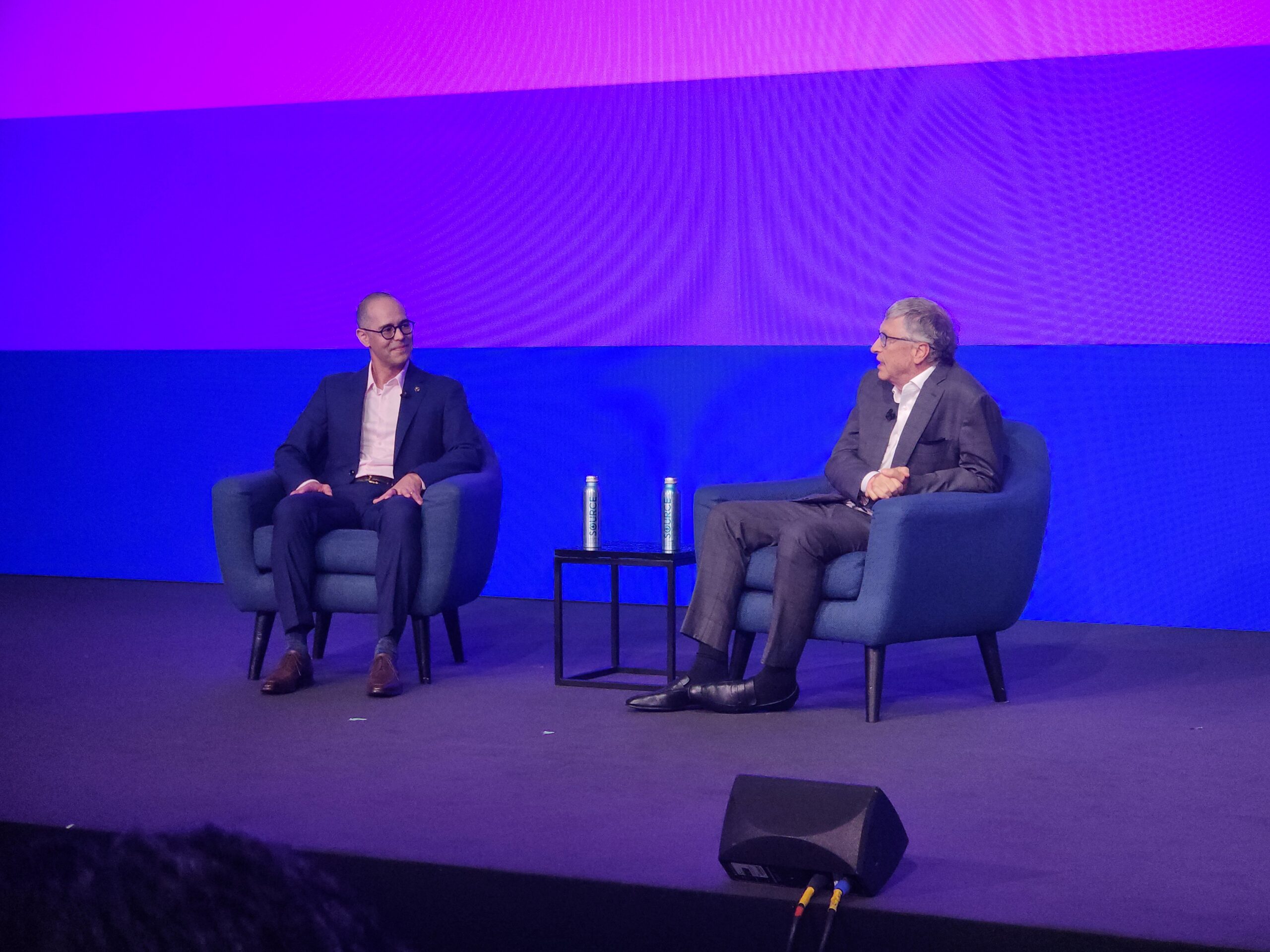 Two men sit on a stage, talking, with a large, colorful video screen behind them.