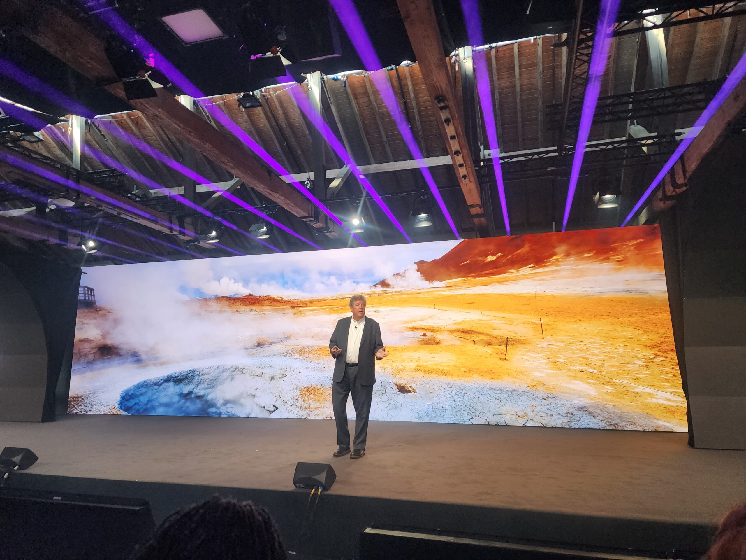 A man stands on a large stage in front of a screen with a colorful picture of a landscape behind him.