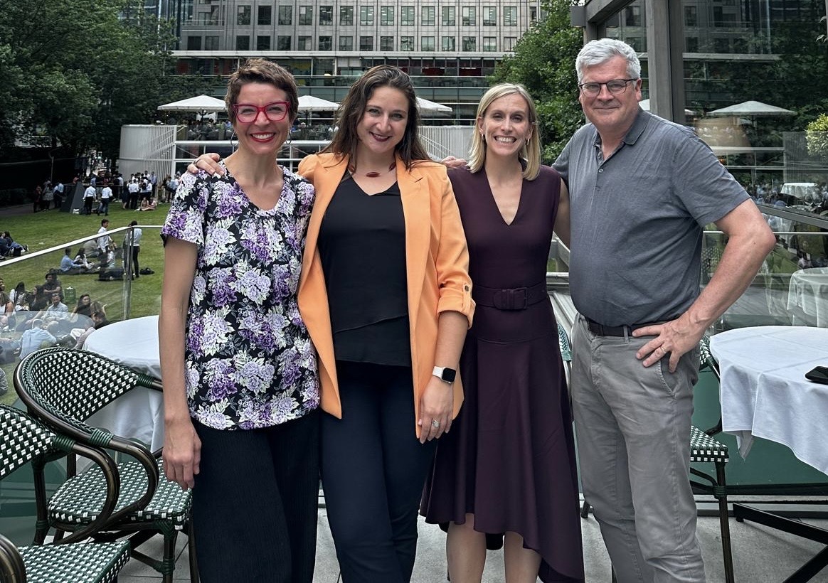 Four people, three women and one man, stand and smile together in front of an outdoor courtyard.