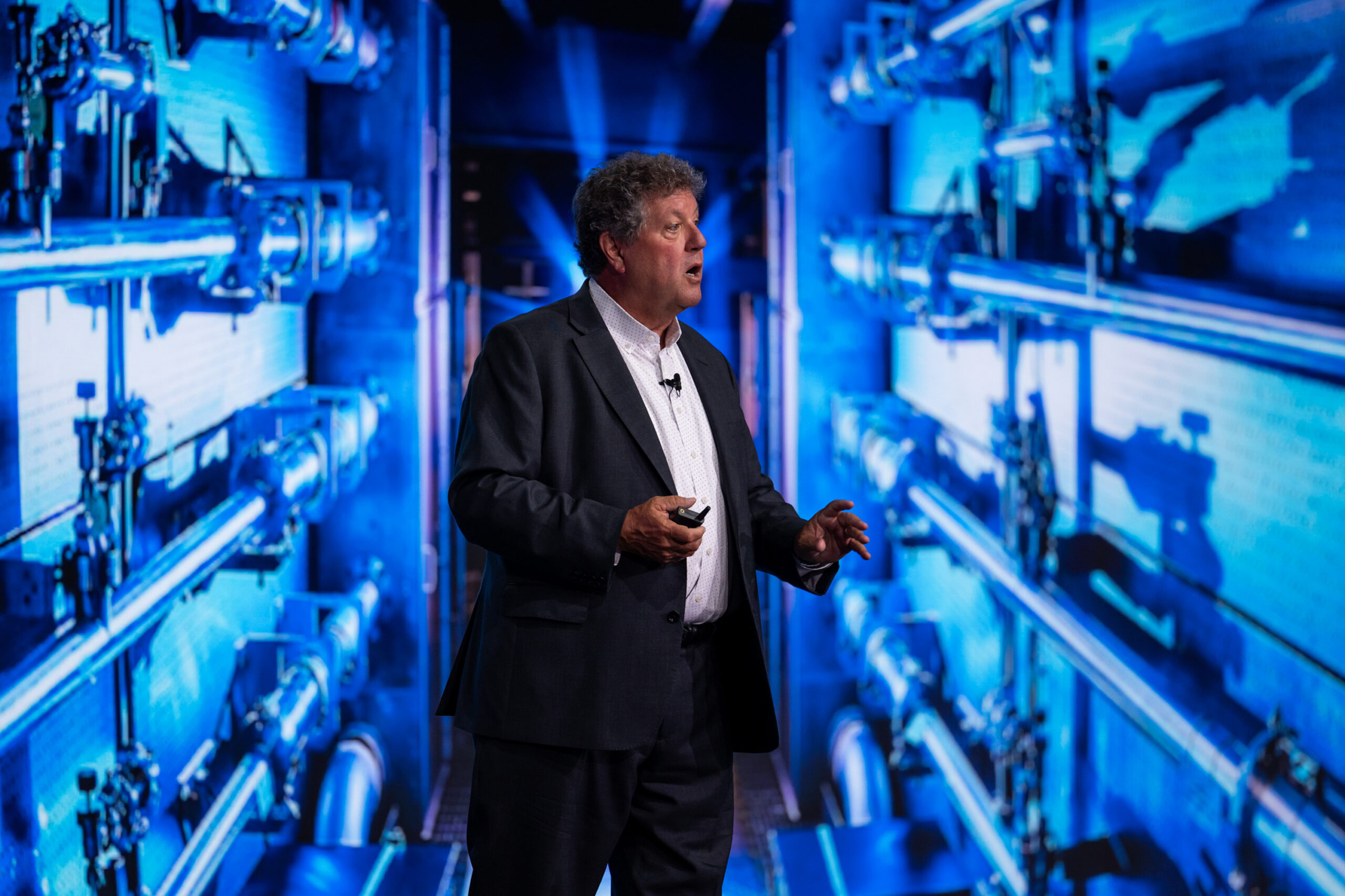 A man gestures while speaking in front of a large video screen showing a bunch of pipes.
