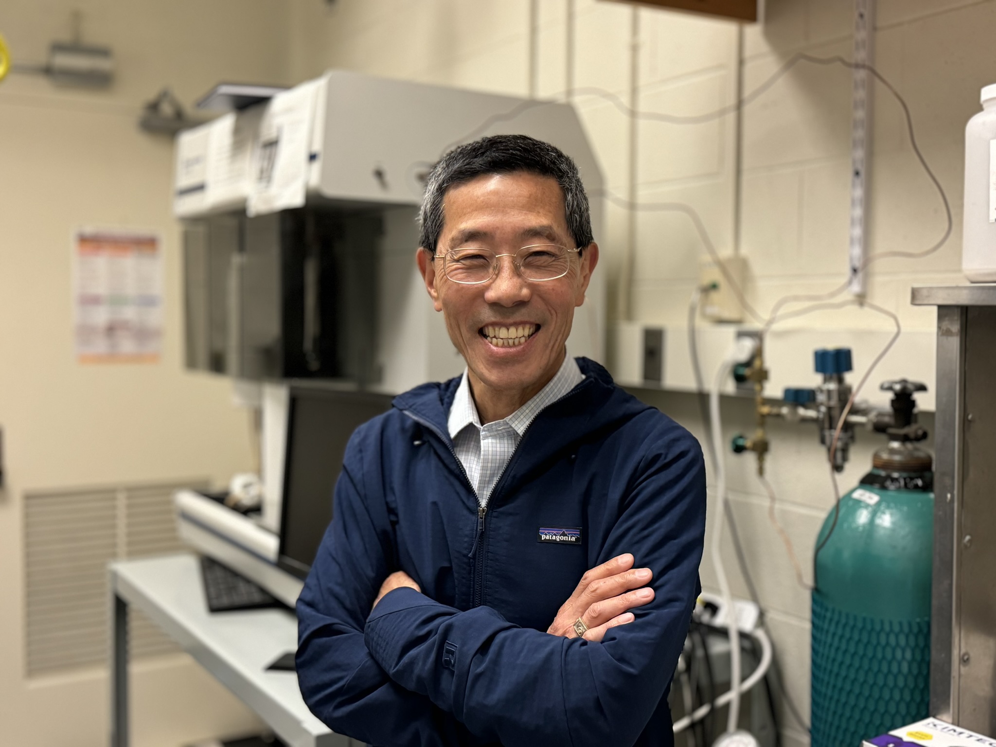 A man stands smiling with his arms crossed in front of scientific laboratory equipment.