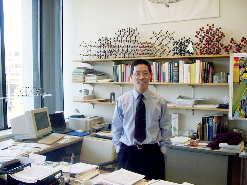 A young man stands with hands in his pockets in an office with stacks of paper everywhere, models of molecules lining a bookshelf, and a clunky early 2000s computer on the desk.