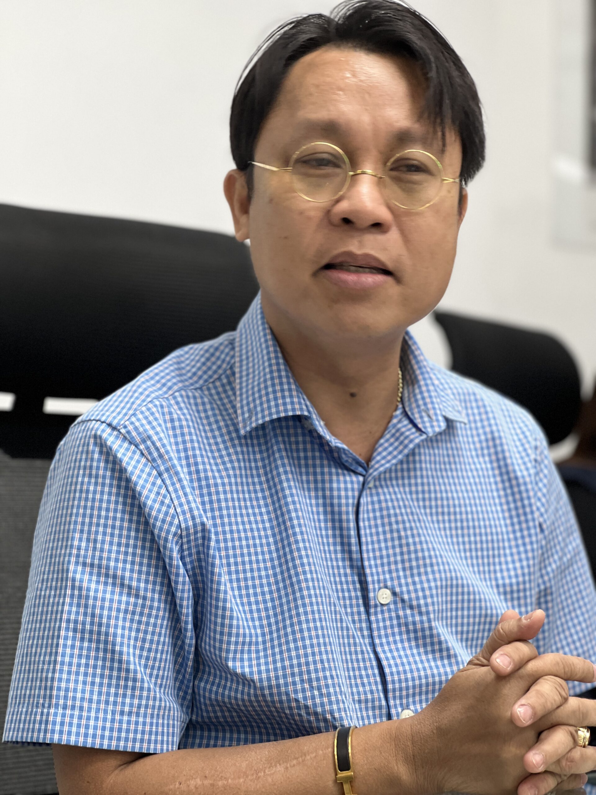A man wearing glasses and a blue shirt speaks with his hands clasped in front of him. 