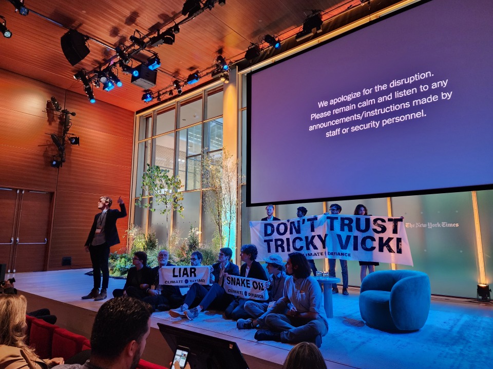 Protesters hold up a sign that says "Don't Trust Tricky Vicki" on stage in front of an audience of people.