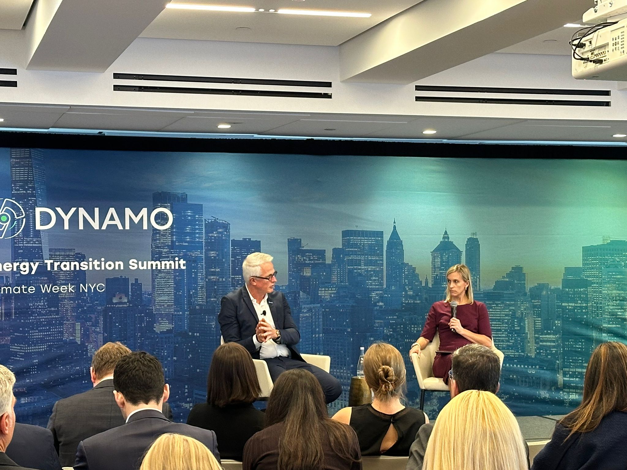 Two people, a man and a woman, are seated and speak in front of a backdrop that says "Dynamo"