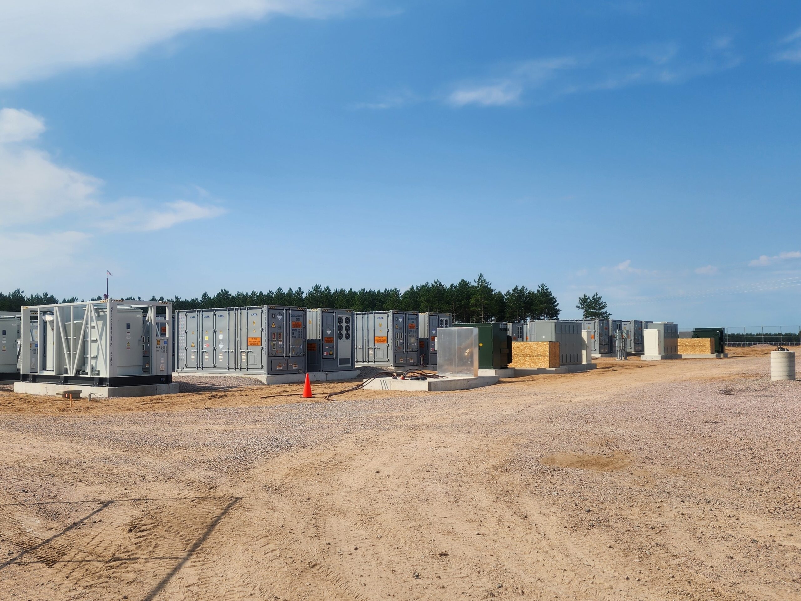 Grey, rectangular banks of batteries the size of storage units sit on a large patch of dirt, with trees in the background.