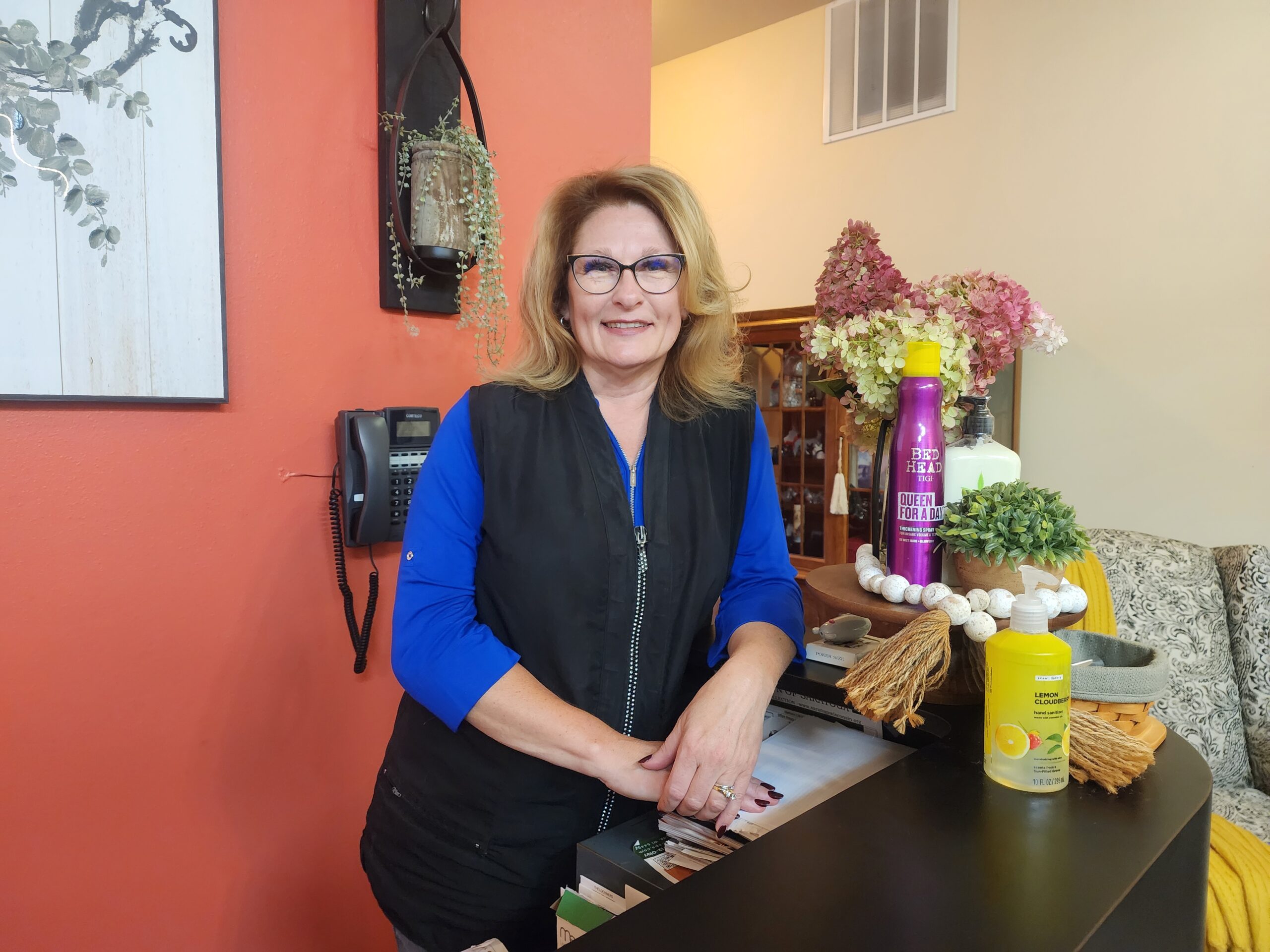 A blonde woman wearing a black vest over a blue long sleeved shirt smiles at the camera. She stands at a reception desk with an orange wall behind her. She wears glasses.