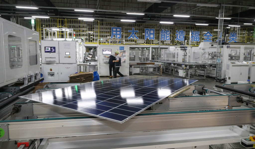 A photo of a solar panel in a manufacturing facility with flourescent lighting and workers in the background and big blue Chinese characters on the wall.