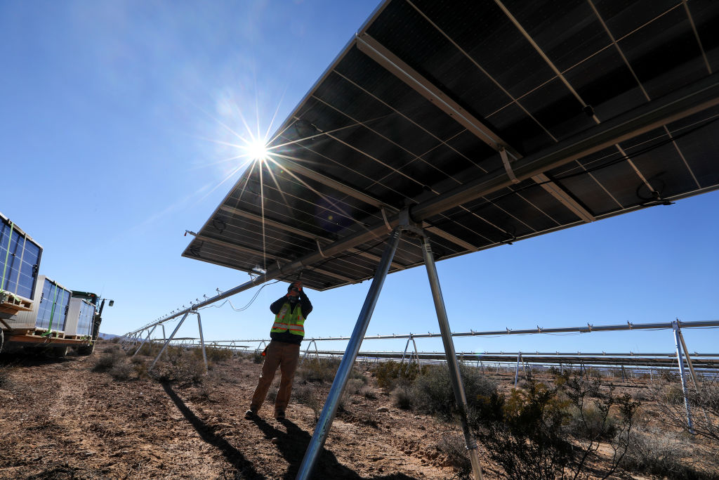Photo of a worker securing a mount underneath a large solar panel, with the sun high in the sky above.