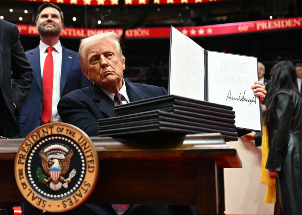 President Donald Trump sits at a desk with the seal of the president of the United States on it and holds up a black folder with two pieces of white paper on it with writing and his large signature in black ink. Vice President JD Vance stands behind him, looking up at the crowd. They are on stage at the Capital One Arena and in the photo you can see red electronic banners that read 