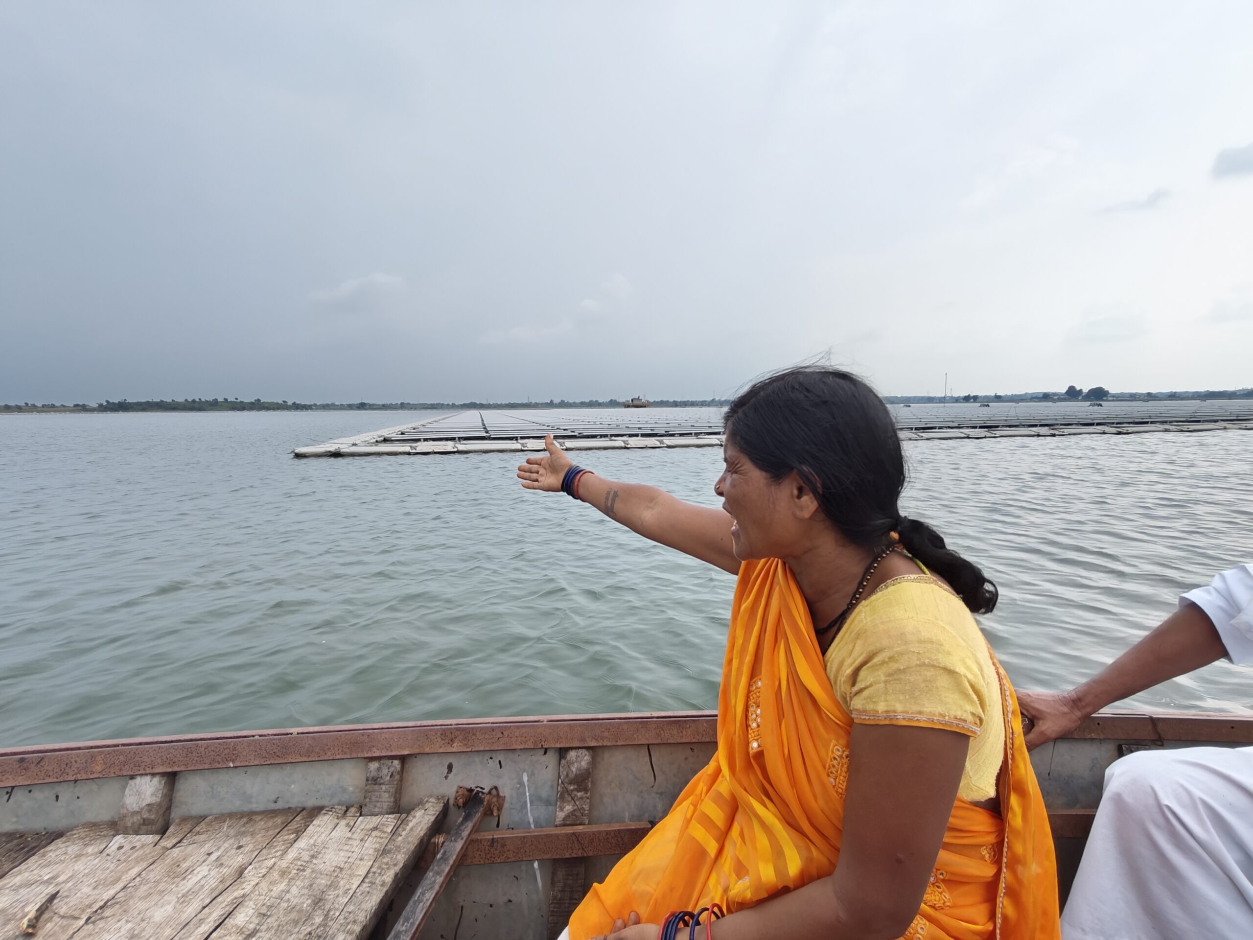Indian woman from rural fishing village wearing an orange sari in a wooden boat on a lake pointing to floating solar panels on the water