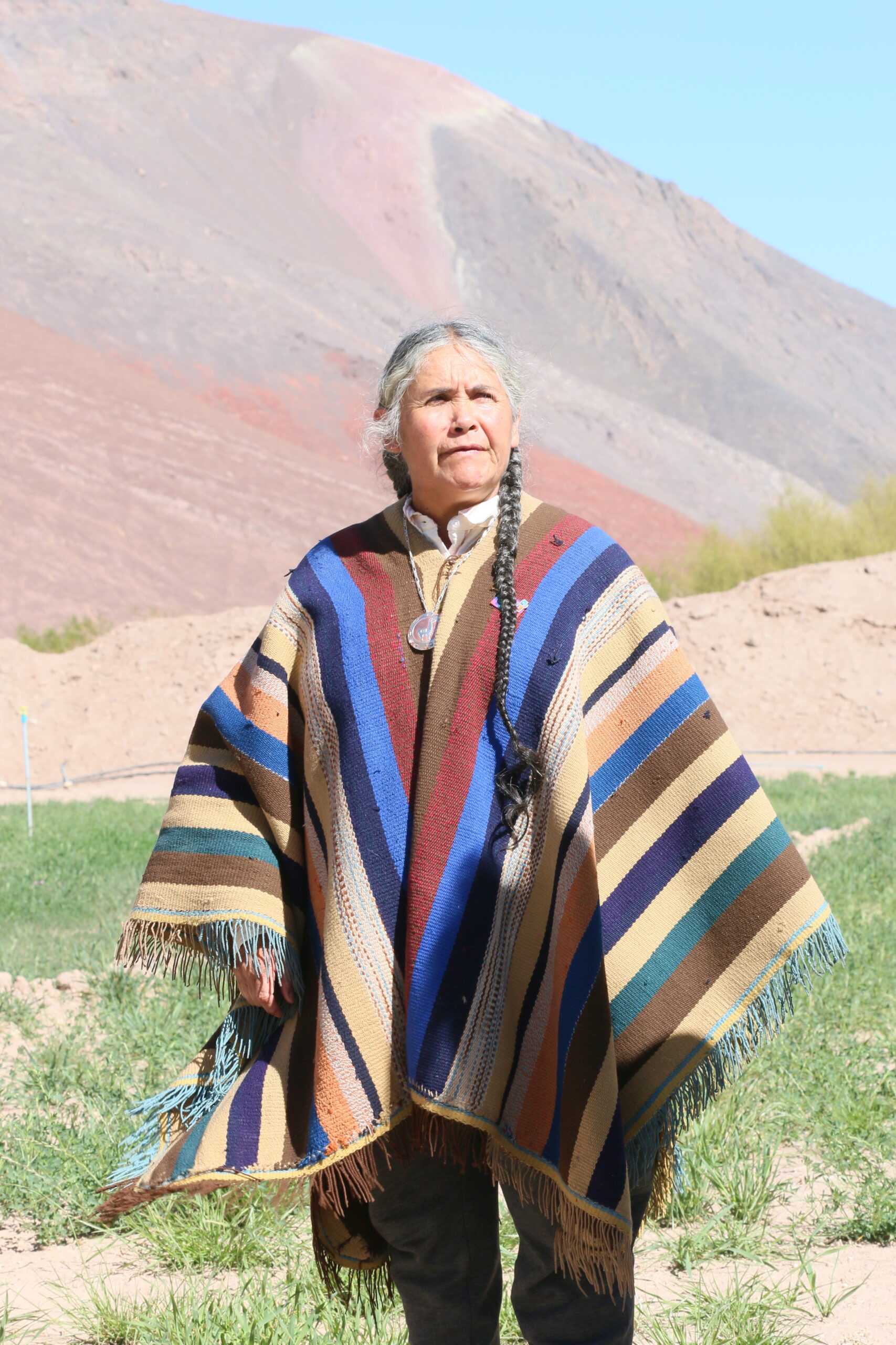 A woman with long grey braids wears a poncho striped with blue, rust red, blue, brown, black, pink and beige, stands in front of a reddish mountainside and a clear blue sky.