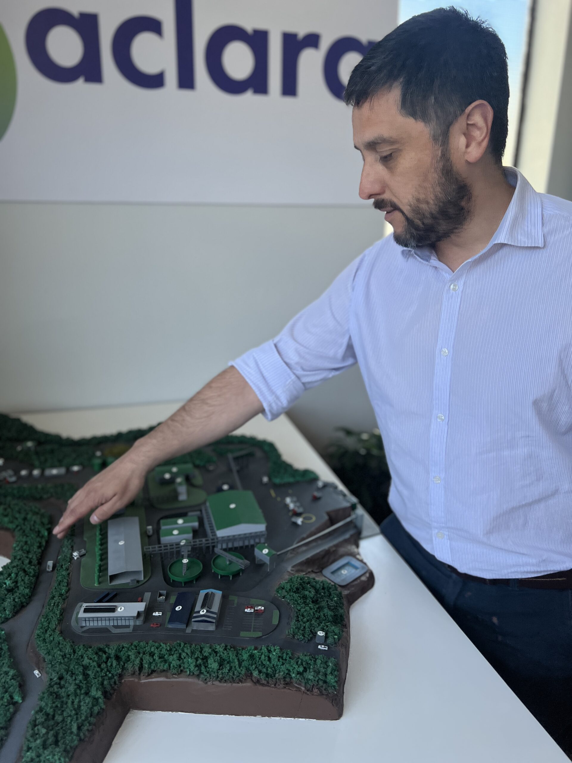 Photo of a man in a light blue shirt pointing to a miniature model of a mining operation on a table in front of him.