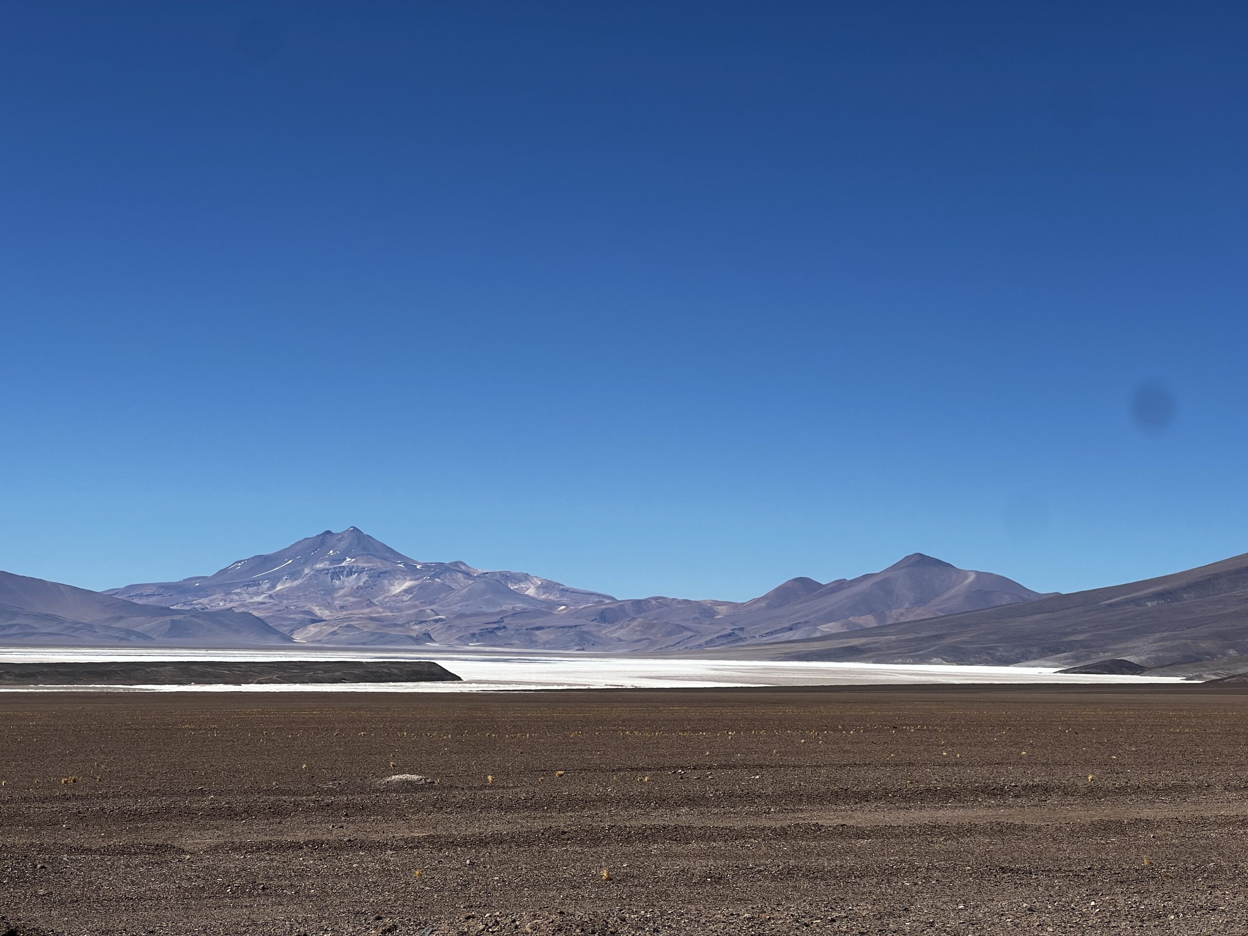 A photograph of a barren looking desert landscape with a white salt lake in the foreground and tall rocky mountains in the background. The landscape has a reddish quality to it and the sky is clear and blue.