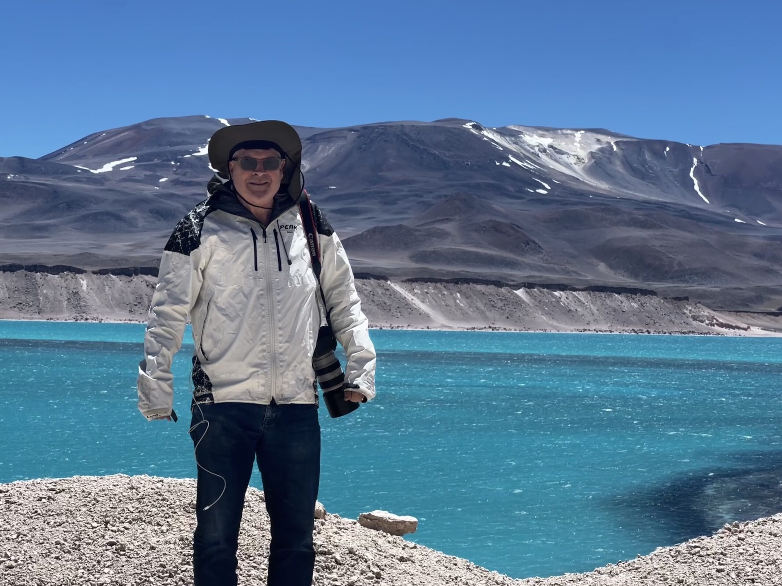 A man in a white windbreaker and a hat and sunglasses, with a large telephoto lensed camera at his side, stands in front of a bright blue lake with a big grey mountain in the background.