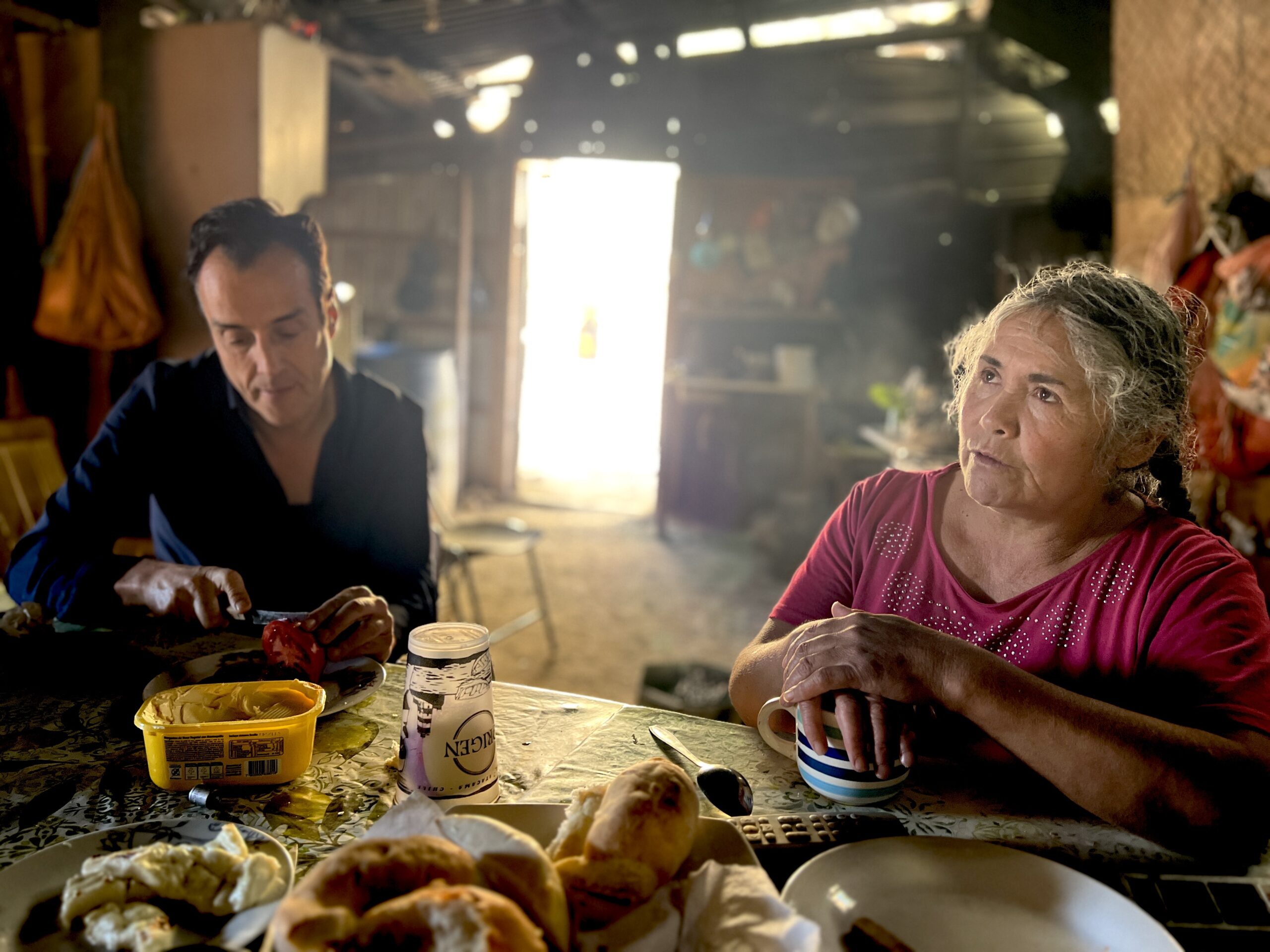 A man (left) and a woman (right) sit around a table laden with food inside a room with a door open to the bright sunshine beyond.