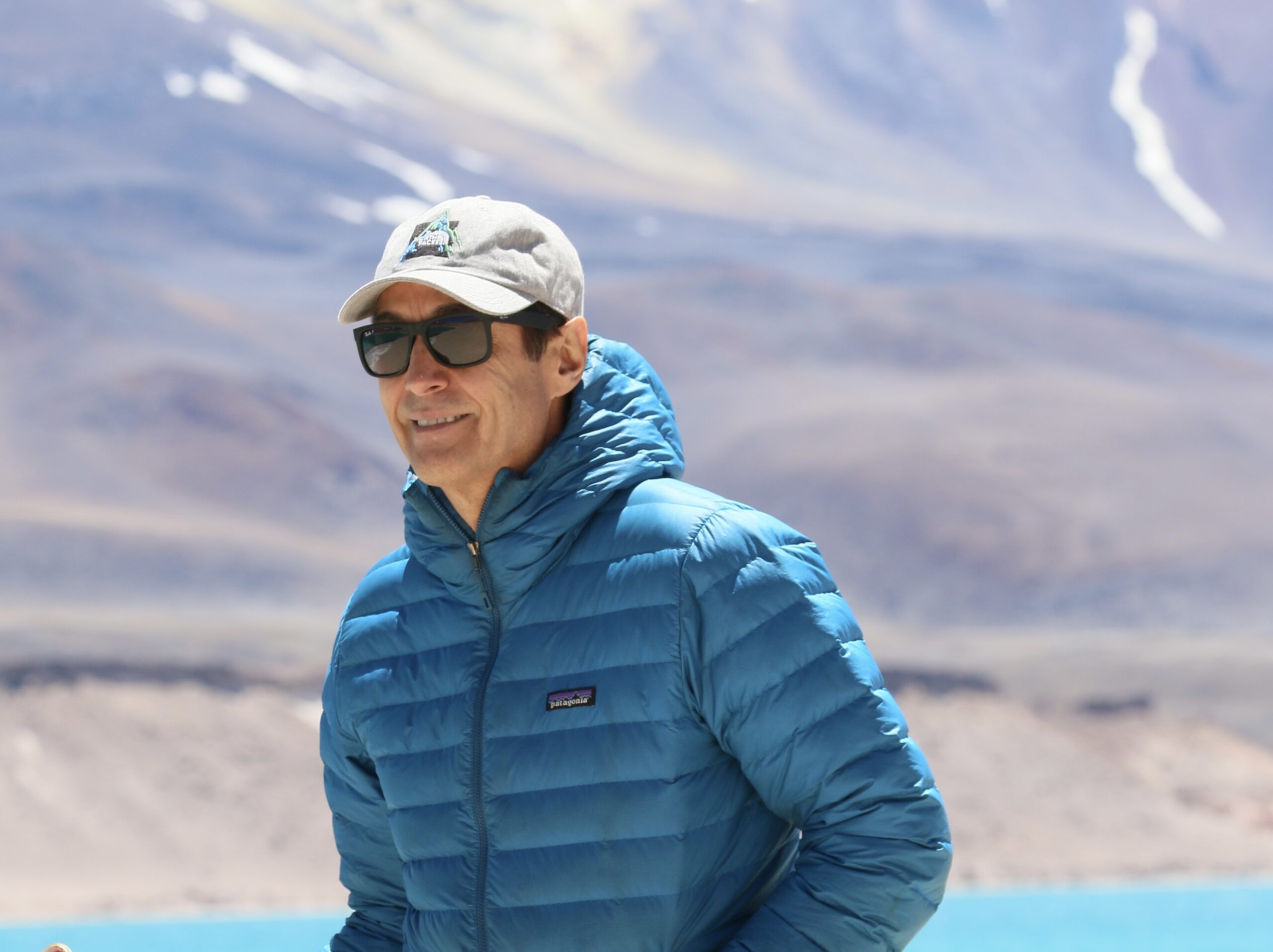 Photo of a man wearing a hat, a blue puffer jacket and sunglasses in front of a mountain and a blue lake.