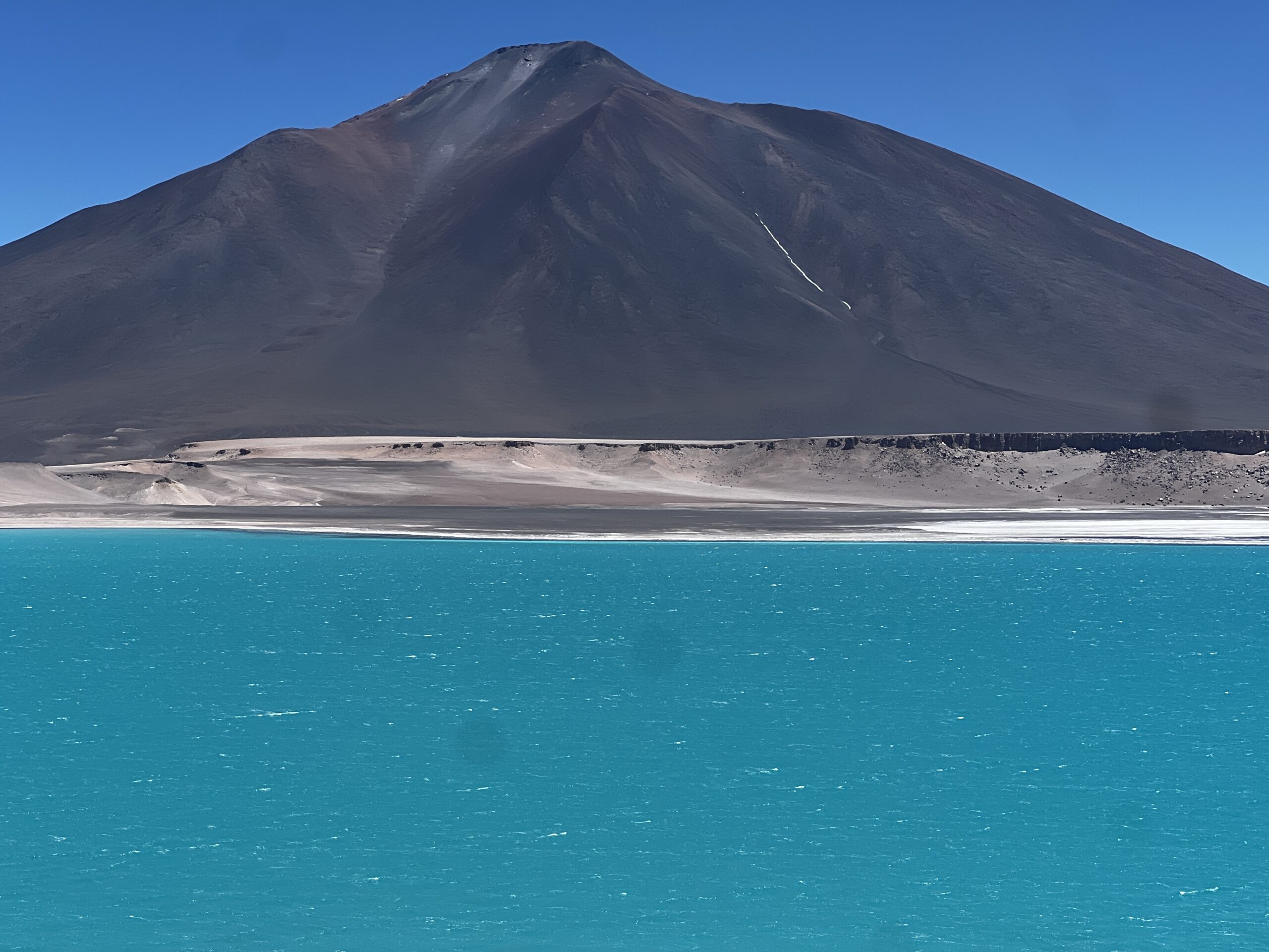 A photograph of a lake with bright blue, almost green, water, and a rocky red and dark gray mountain behind it.
