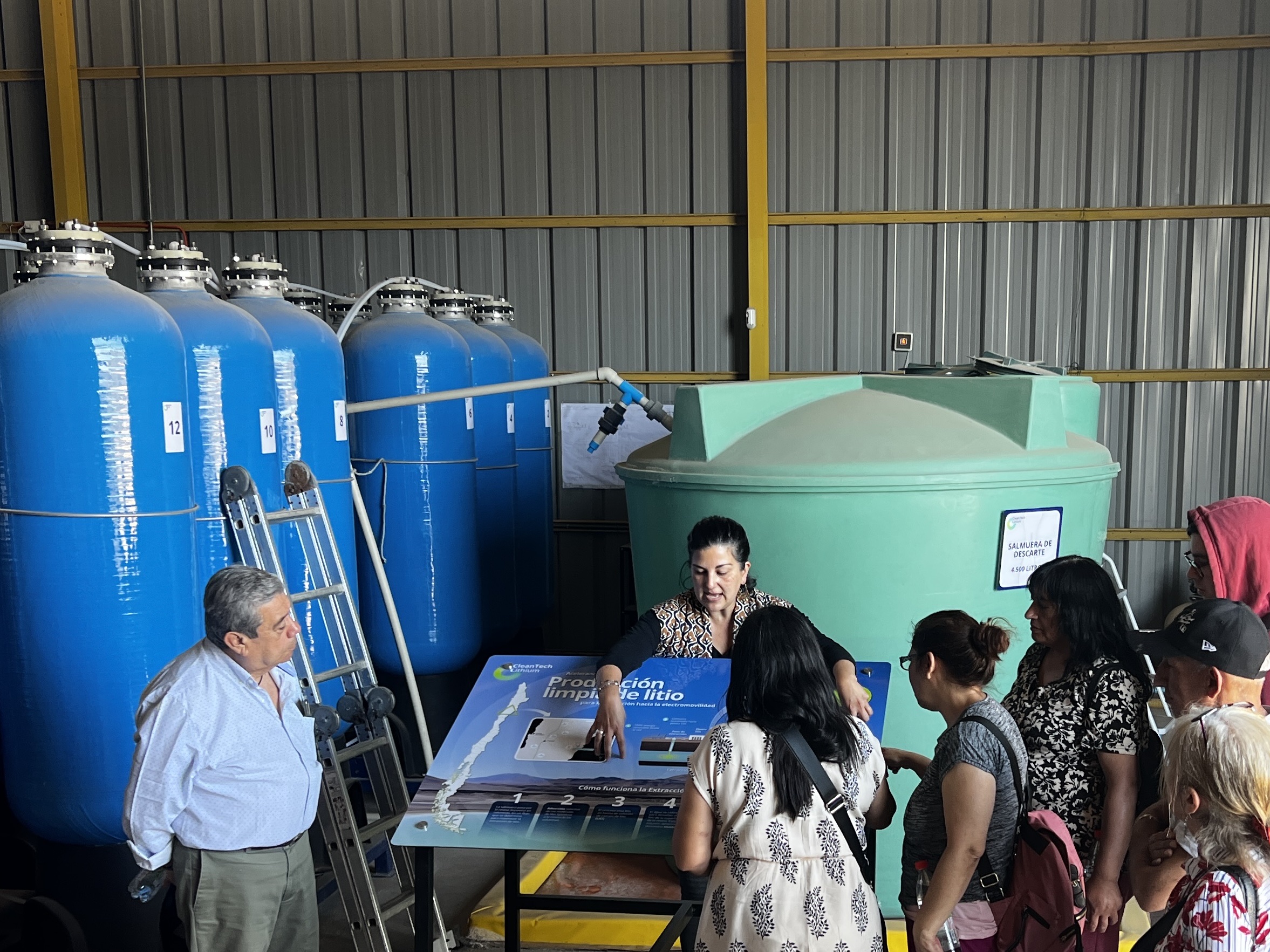 Photo of a group of people gathered around a board demonstrating a lithium extraction process (the board is written in Spanish) and surrounded by blue and green tanks. A woman stands behind the board, gesturing to it and talking.