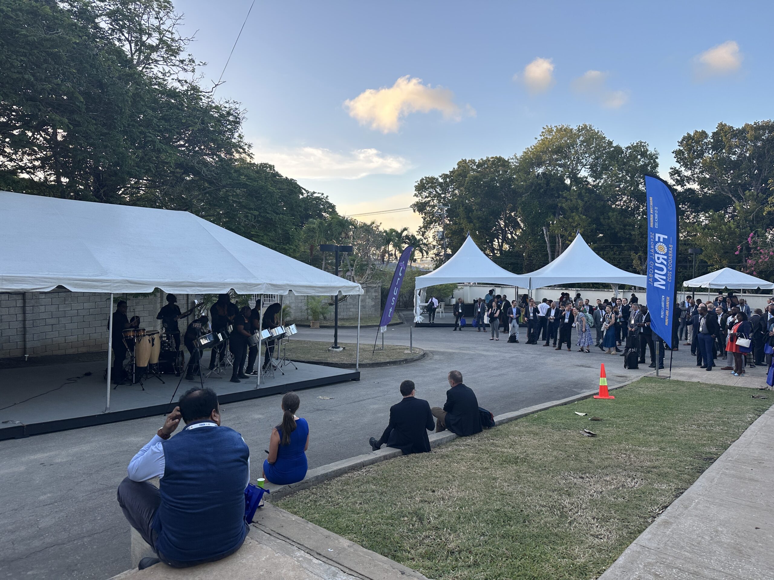 A photo of people playing steelpans under a white tent, with a large group of people mingling and listening under other white tents nearby. The event is happening outdoors under a blue sky near dusk.