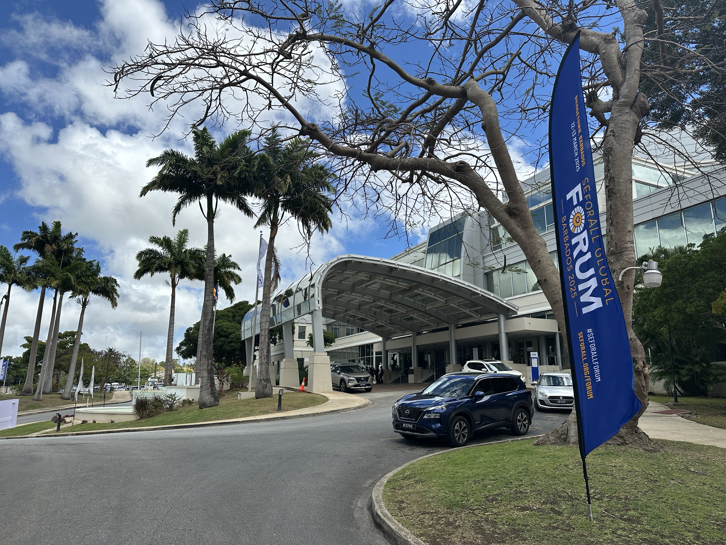 A phot of the outside of a large glass building with palm trees out front, a blue sky, and a flag in front of the building that reads 