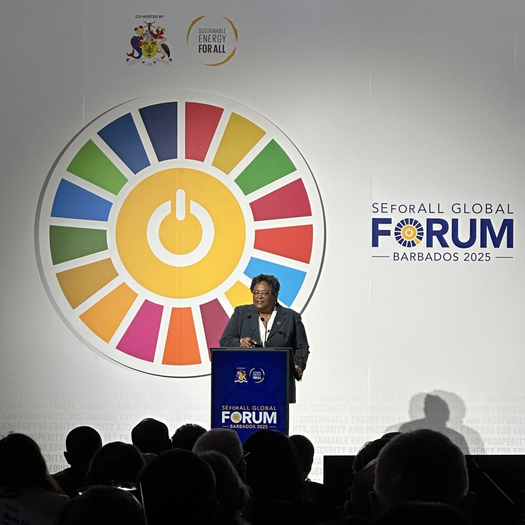 A woman stands at a podium on stage and speaks to a large audience. The backdrop is white with a colorful pinwheel with a power button in its center and the words "SEforALL Global Forum Barbados 2025" next to it.