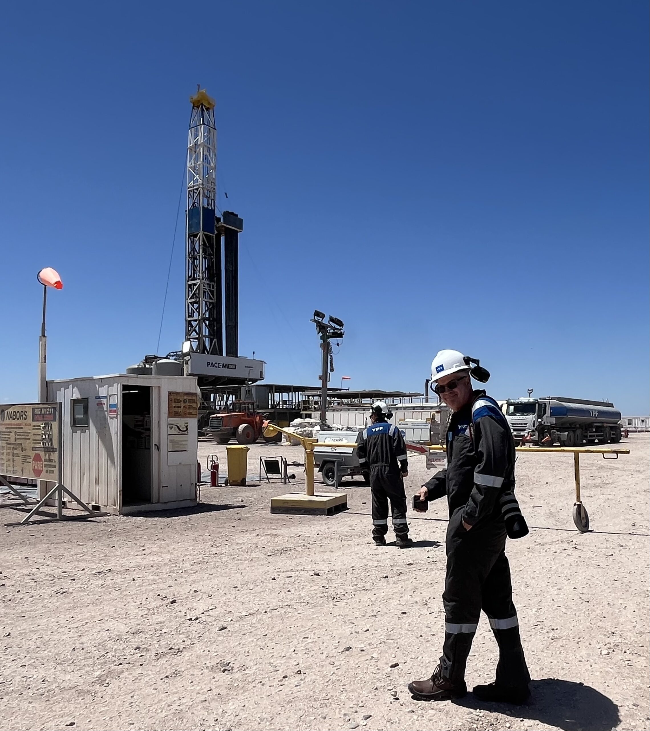 In this photo, a man wearing a black industrial jumpsuit with neon trimming and a hard hat grins at the camera as he walks toward an industrial site on a barren-looking landscape.