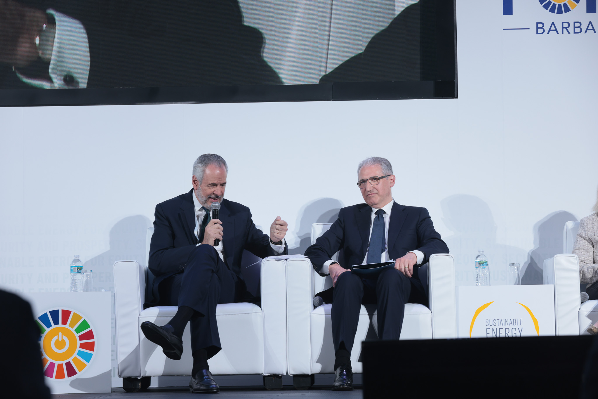 Two men sit on stage in front of a white background; the man on the left holds a microphone and is gesturing while he talks. Both men are wearing suits.