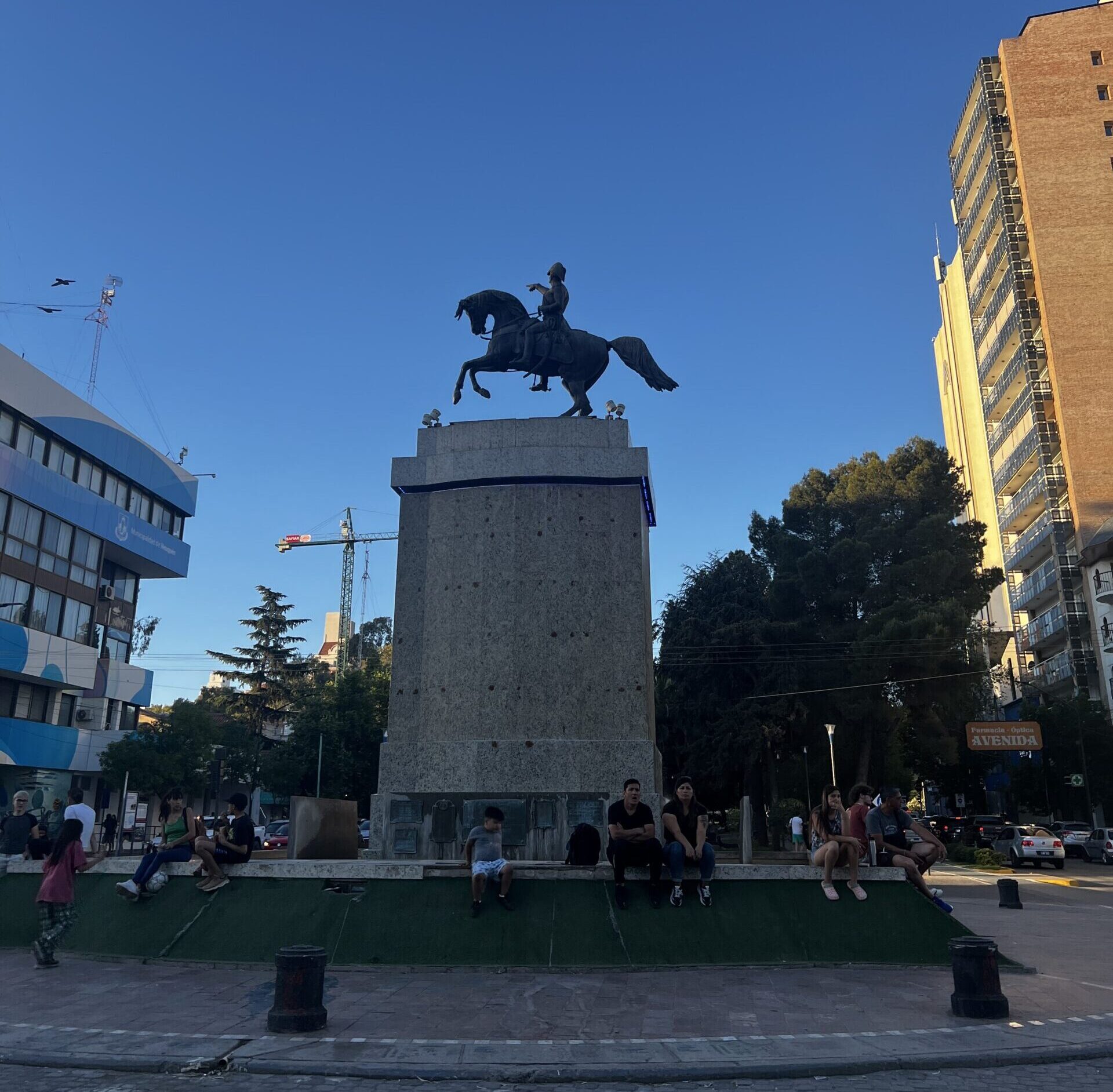 A photo of a large concrete monument with a mounted rider on a horse on top. The horse statue has two hooves on the ground. People sit around the base of the monument, buildings loom on either side and a crane is visible in the background.