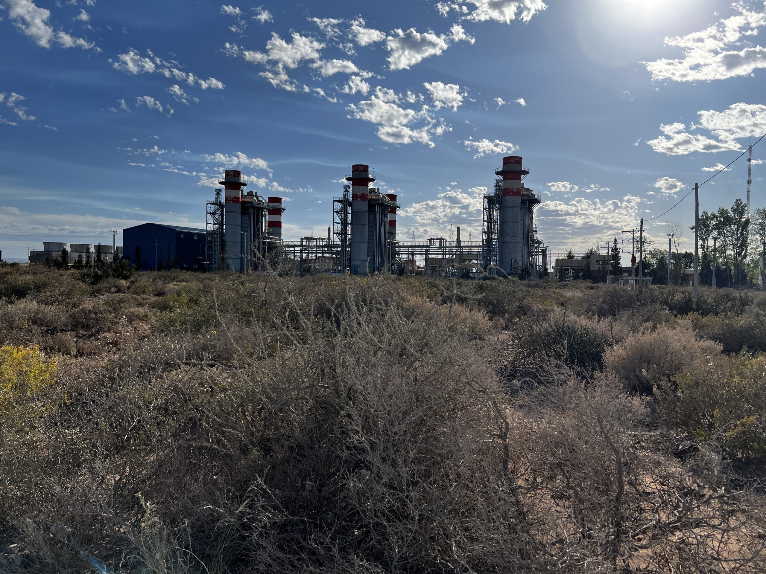 A photo of an industrial facility under a blue sky dotted with white clouds. In the foreground, there's brush and small, dry-looking plants.