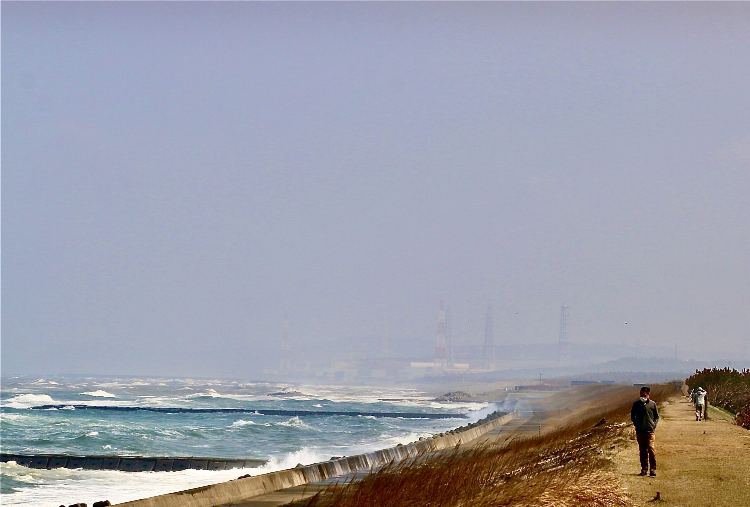 A photo of a beach and a blustery sea, with a large industrial facility visible behind some haze in the background.