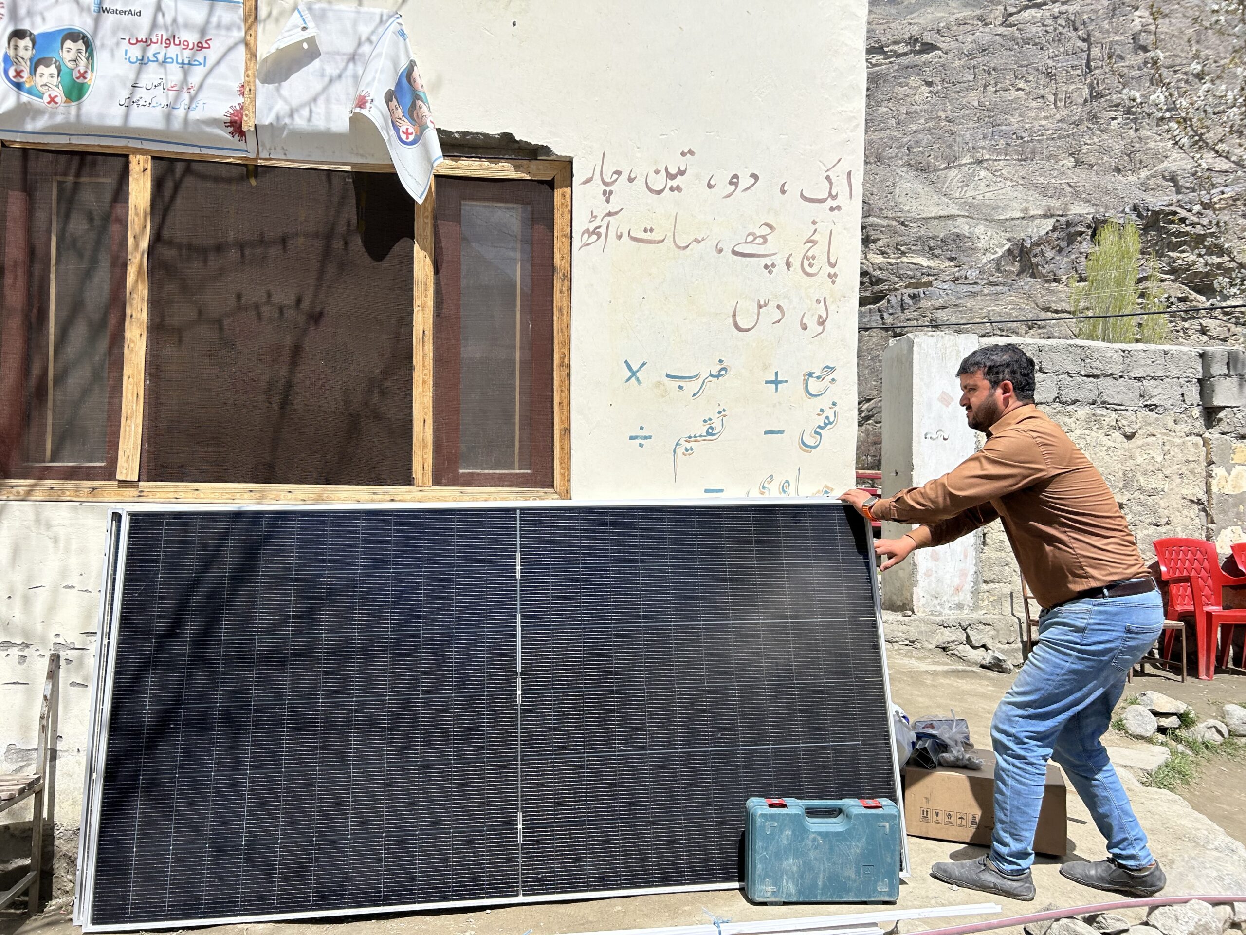 A man in a brown shirt adjusts a solar panel in front of a yellow outdoor wall with writing on it in Urdu.