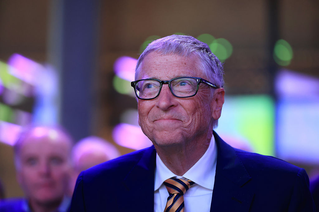 A photo of an older man wearing glasses, looking up hopefully with a little smile on his face. He wears a suit and tie and the light around him is purple. There are other people and screens out of focus in the background.