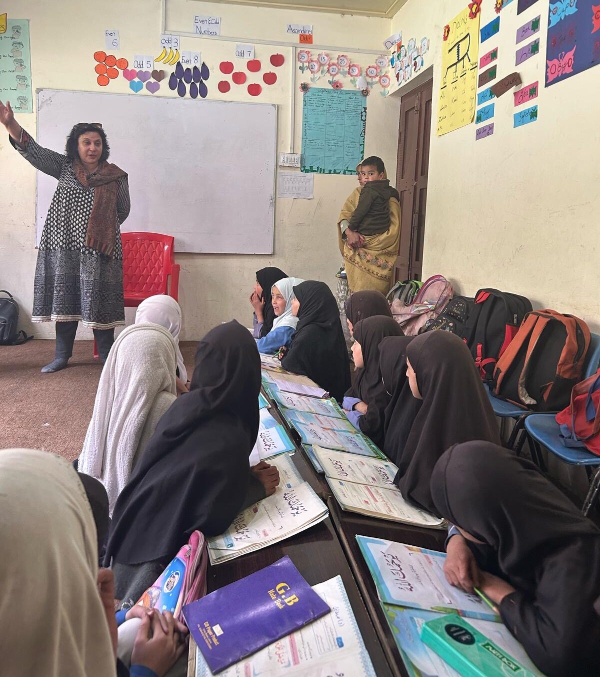 A woman stands at the front of a classroom of girls watching her gesture in front of a white board.